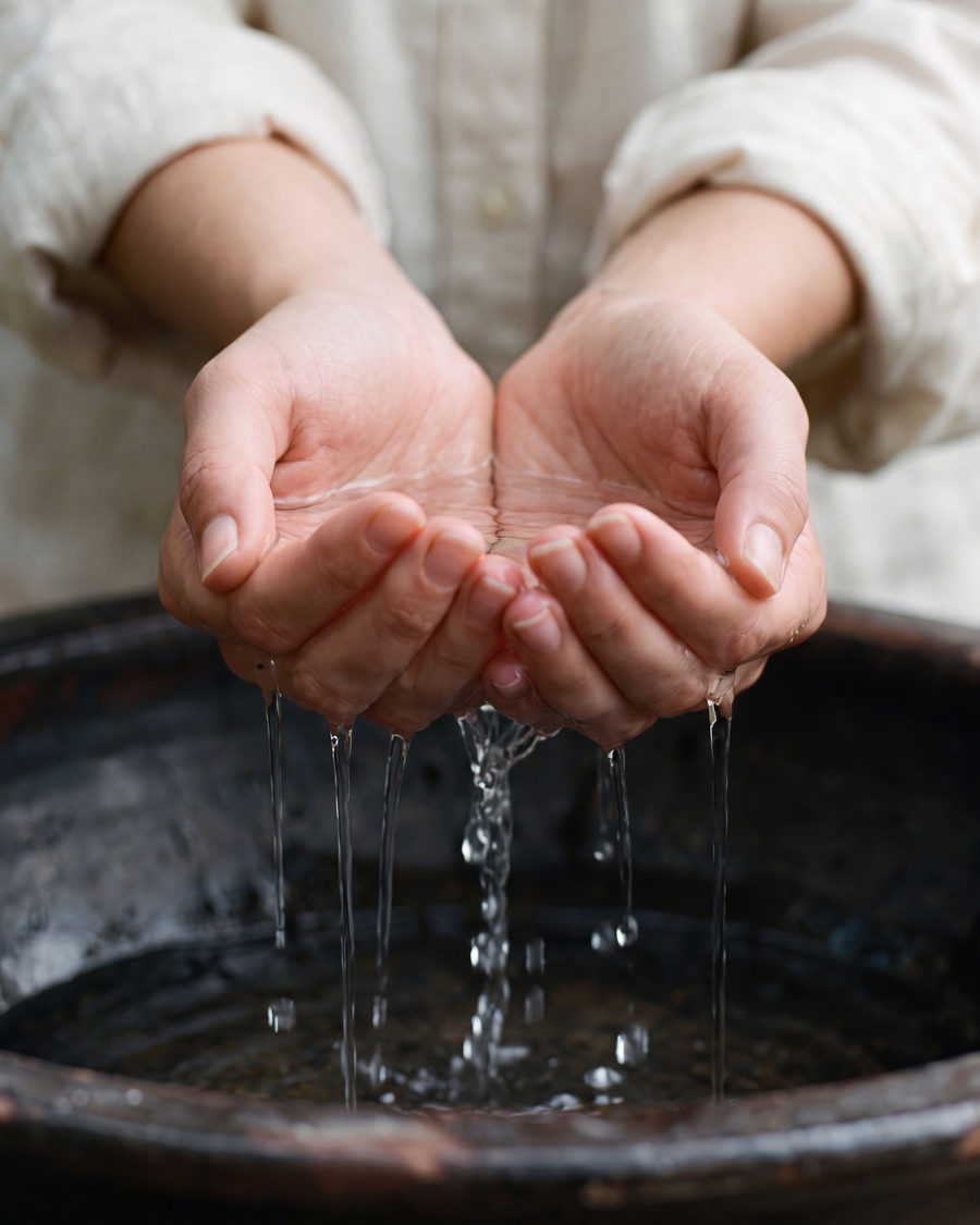 Zwei Hände schöpfen klares Wasser aus einem dunklen Steinbecken; das Wasser läuft zwischen den Fingern zurück
