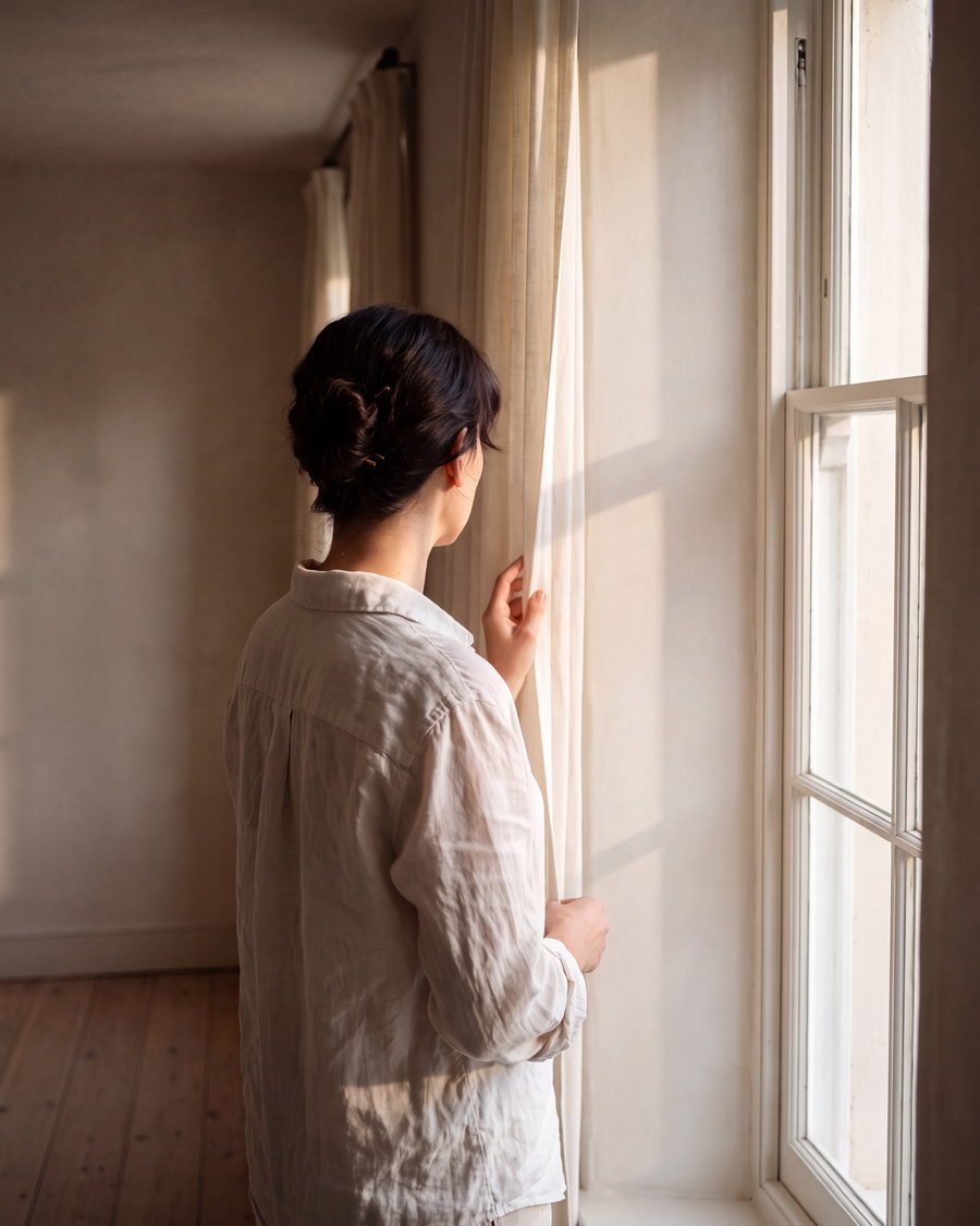 Eine Frau von hinten an einem hohen Fenster, das Haar hochgesteckt, Morgenlicht auf der Schulter